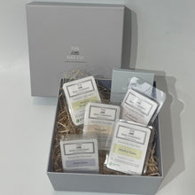 A gift box open to reveal multiple packaged wax melts with labels, accompanied by a bed of decorative straw. An additional pack of tea lights is also visible.