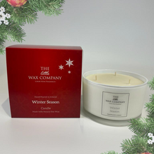 A wax candle with a label 'Winter Season' from The Little Wax Company, placed next to its red packaging box with 'Christmas Candle' text, decorated with artificial holly and pine needles around.