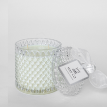 A large diamond-shaped white candle in a clear cut glass vessel with embossed geometric patterns, accompanied by a decorative lid and a brand tag.