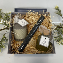 Gift set with candle, bottle, and black tool in a box with straw, surrounded by greenery.