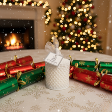 White textured candle with a decorative label on a table with Christmas crackers and a fireplace in the background.