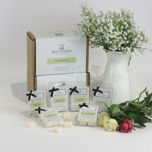 A collection of wax melt cubes from The Little Wax Company, displayed in front of a box with a 6 pack indication. The cubes are labeled with different fragrance names and are presented in clear packaging with black bows.