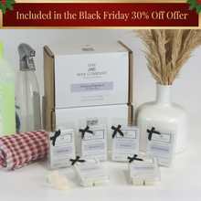A collection of wax melt cubes from The Wax Company, displayed in front of a box with cleaning fragrances. The cubes are labeled with different scent names and are presented with a vase of dried plants and cleaning supplies in the background.