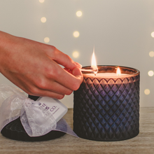 A large navy blue candle in a cut glass vessel with diamond embossing, being lit by a person's hand. The candle is placed on a wooden surface, and there is a decorative bow on top of the vessel.